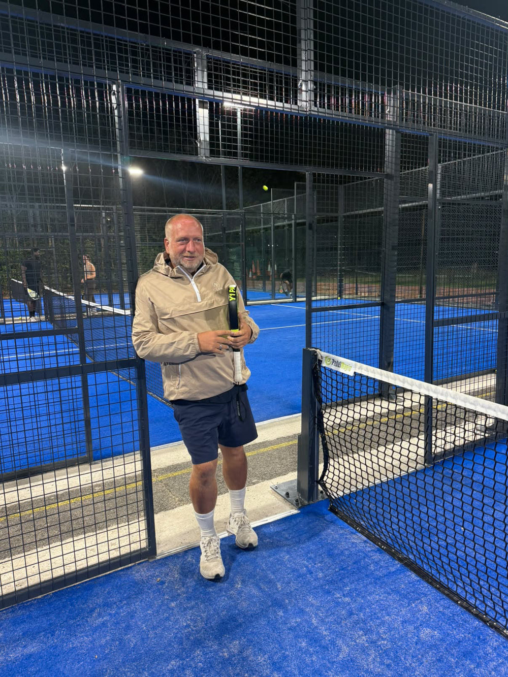 Gary Lowe wearing an AP Club shell beside a floodlit padel court