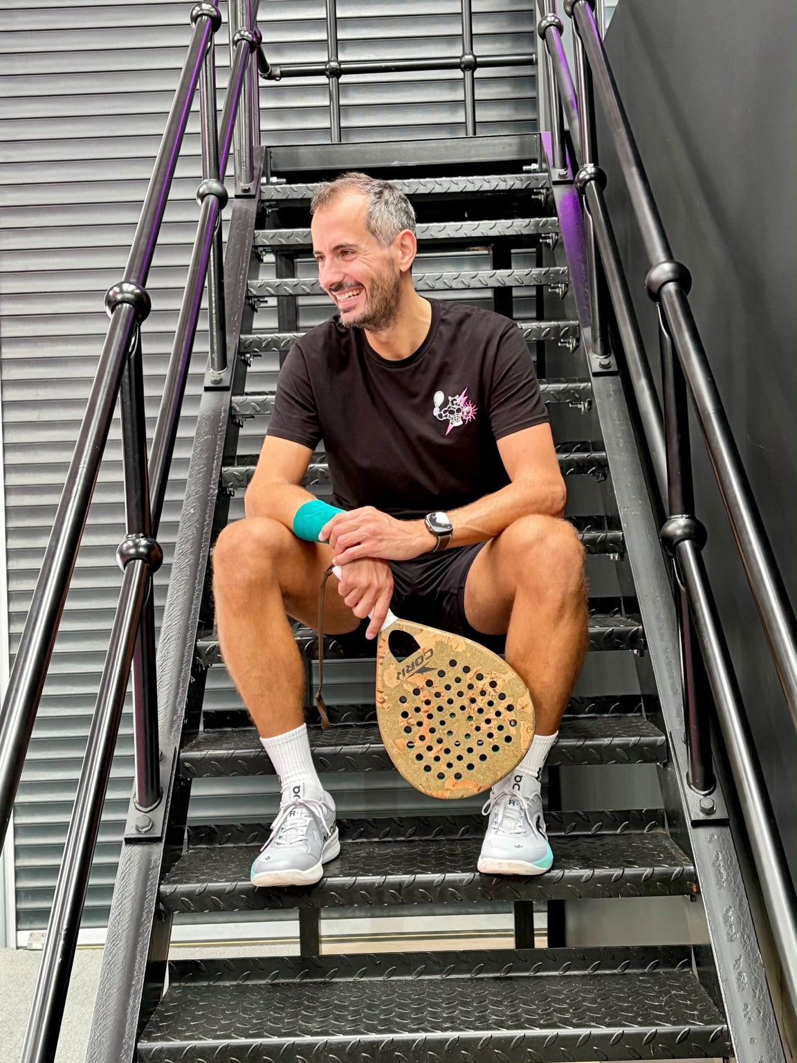 Ross Dyke of Ross Plays Padel seated on metal stairs holding a padel racket