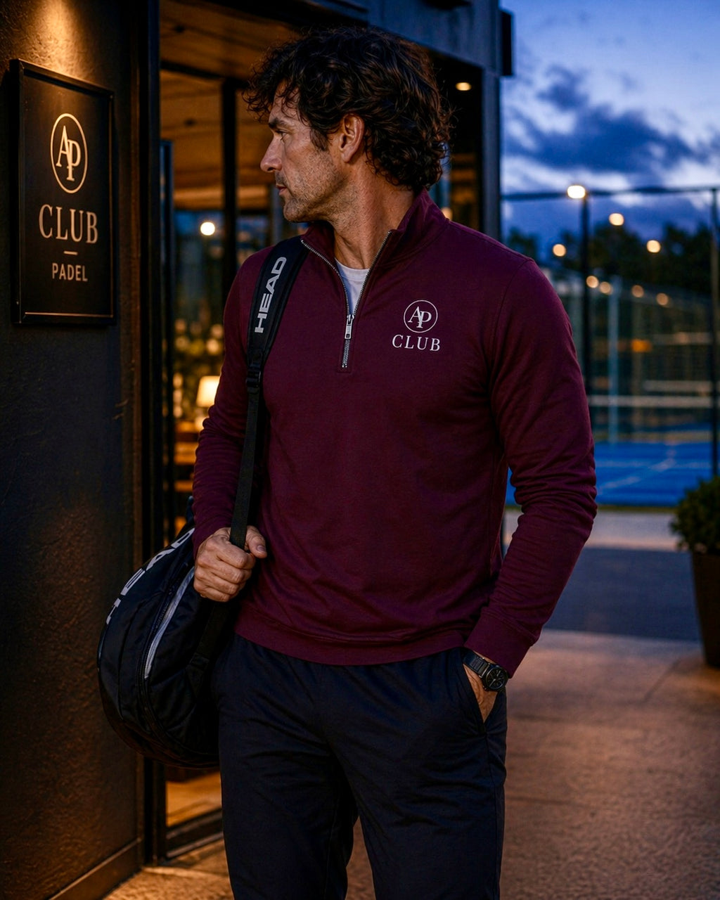 Man in maroon AP Club Quarter Zip with club logo holding a bag, standing outside a building with 'AP Club Padel' sign.