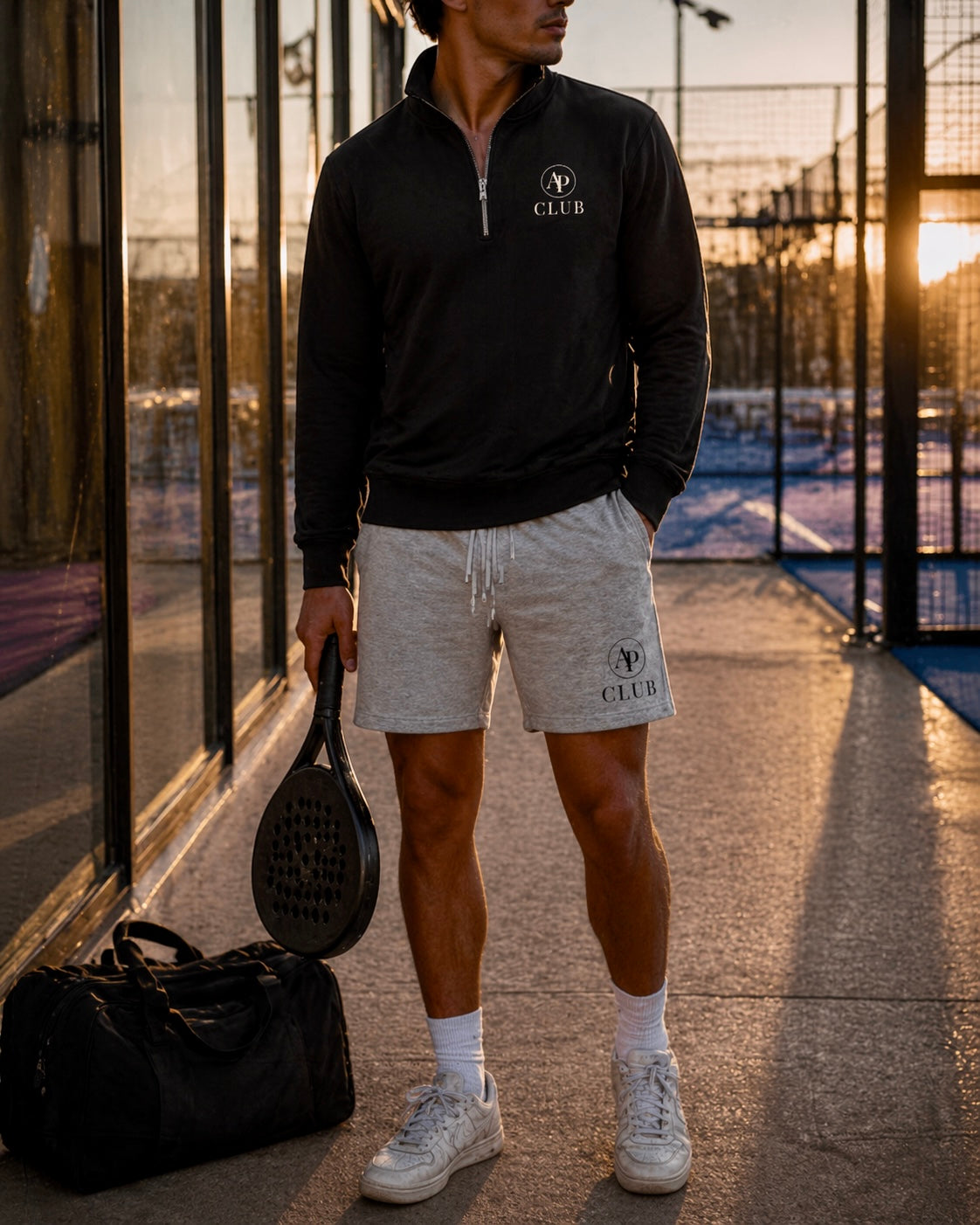 Person wearing a black AP Club Quarter Zip jacket and grey AP Club shorts with a club logo, holding a paddle on a court.