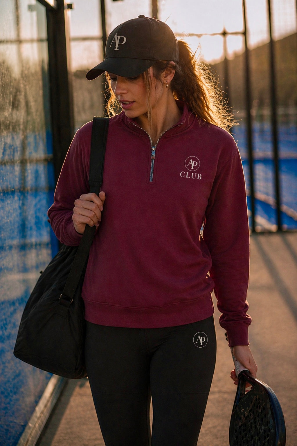 Woman in maroon AP Club Quarter Zip and black AP Club cap with logo, holding a bag and padel racket on a court.