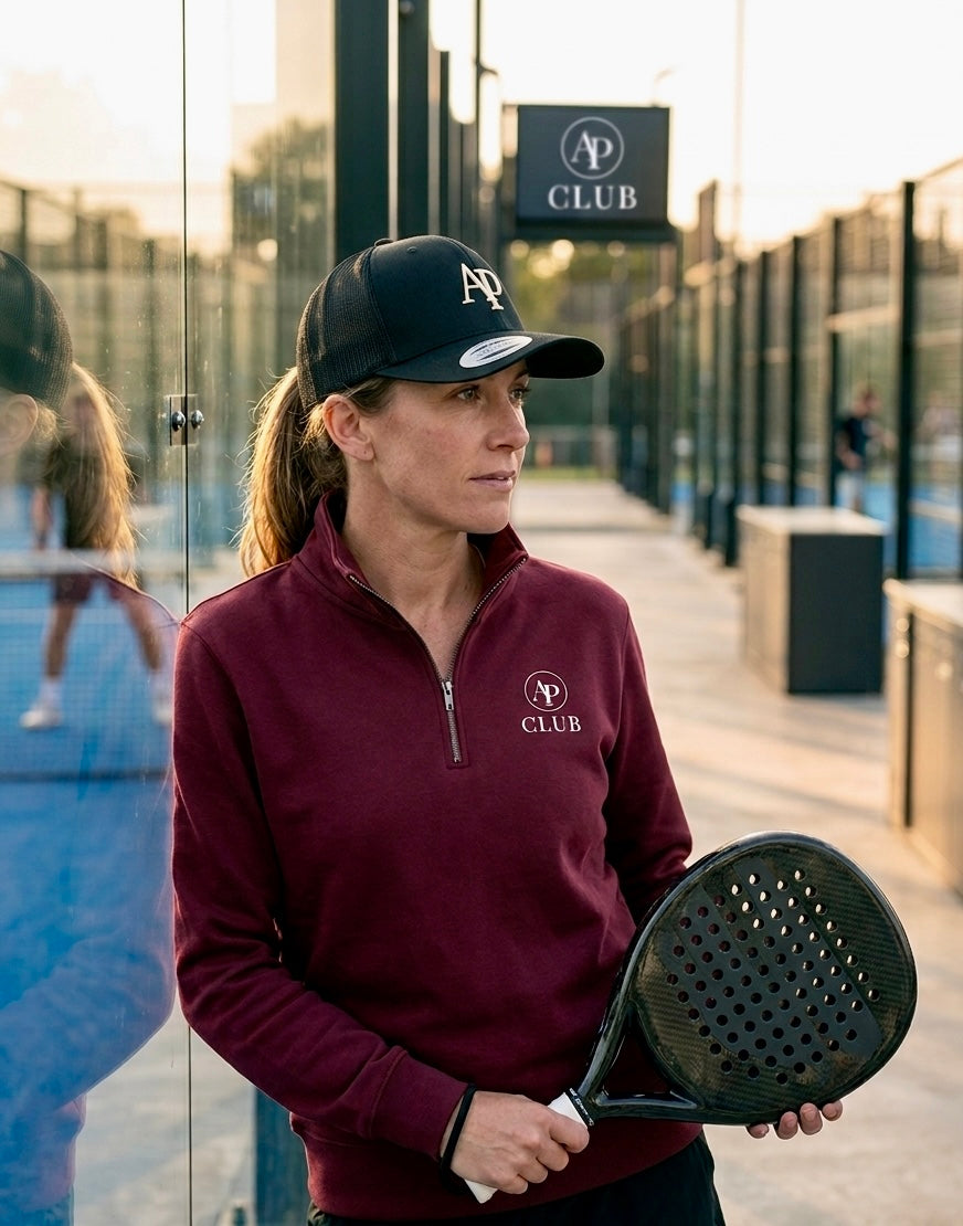 Woman holding a paddle near a glass padel court wearing AP Club Quarter Zip with 'AP Club' branding and black AP Club cap