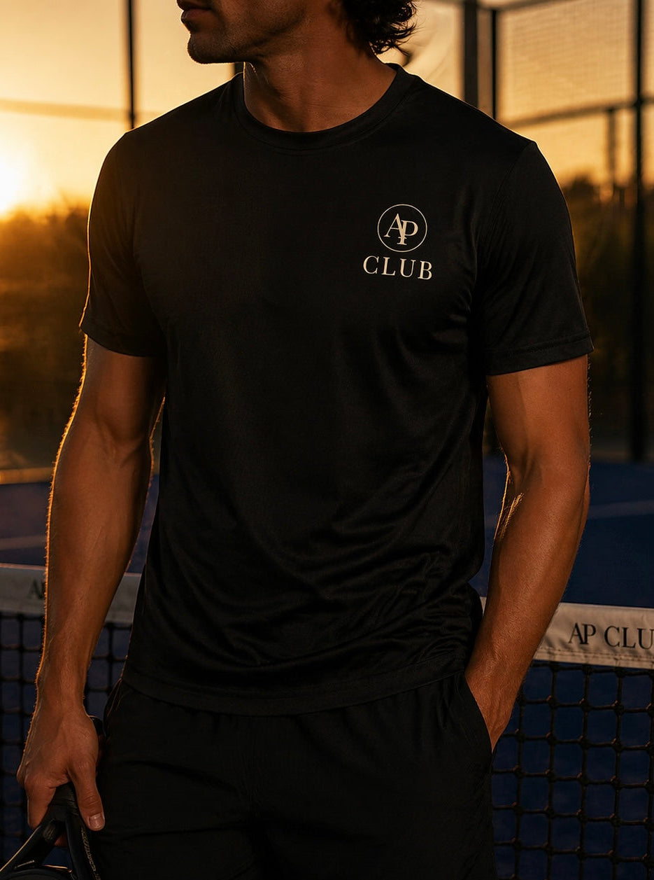Person wearing a black AP Club play t-shirt with a logo and text, holding a padel racket on a padel court at sunset.