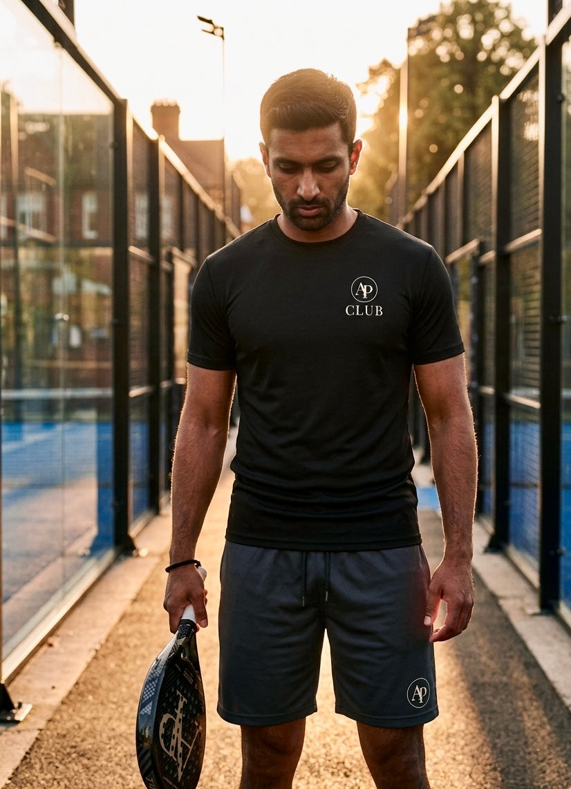 Man holding a paddle on a padel court wearing a black aP Club play t-shirt and dark AP Club court shorts.