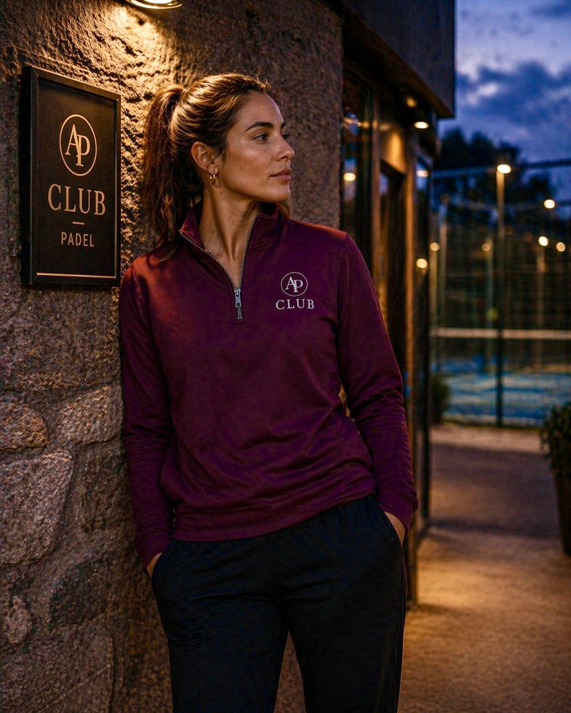 Woman wearing a maroon AP Club Quarter Zip  with a logo, standing outside a padel building at night.