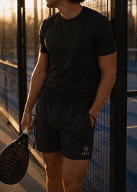 Man in black athletic AP Club Court shorts holding a paddle on a padel court at sunset.