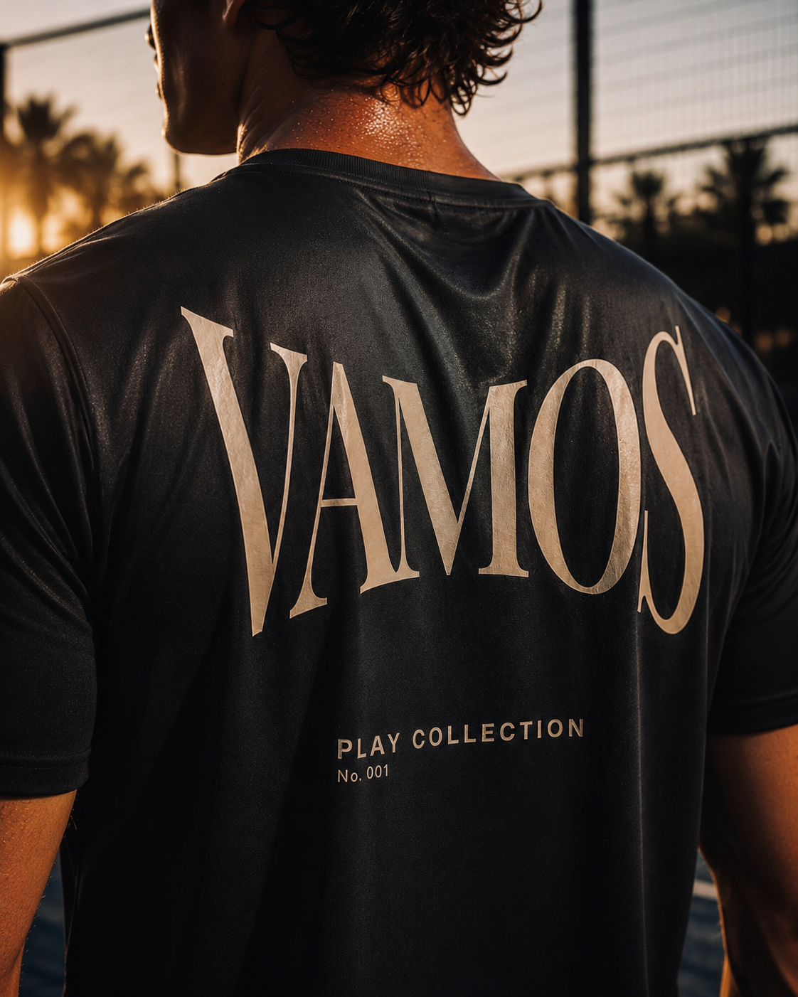 A man viewed from behind wearing a black AP Club Vamos Tee athletic t-shirt with 'VAMOS PLAY COLLECTION No. 001' printed in beige on the back, set against a warm sunset background.