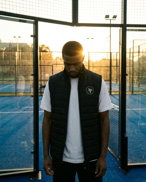 Man wearing a black AP Club gilet over a white shirt with a logo, standing on a blue padel court.