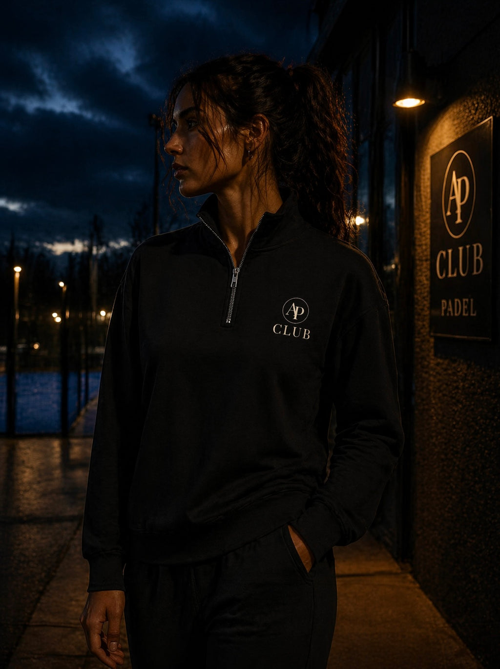 Person wearing a black AP Club Quarter Zip with a logo at night, standing near a padel building with a sign.
