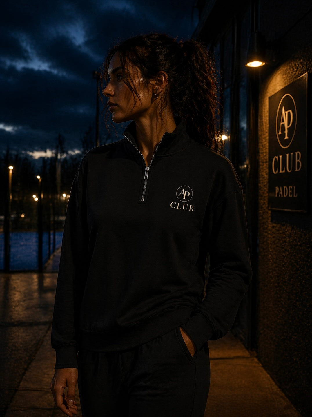 Person wearing a black AP Club Quarter Zip with a logo at night, standing near a padel building with a sign.