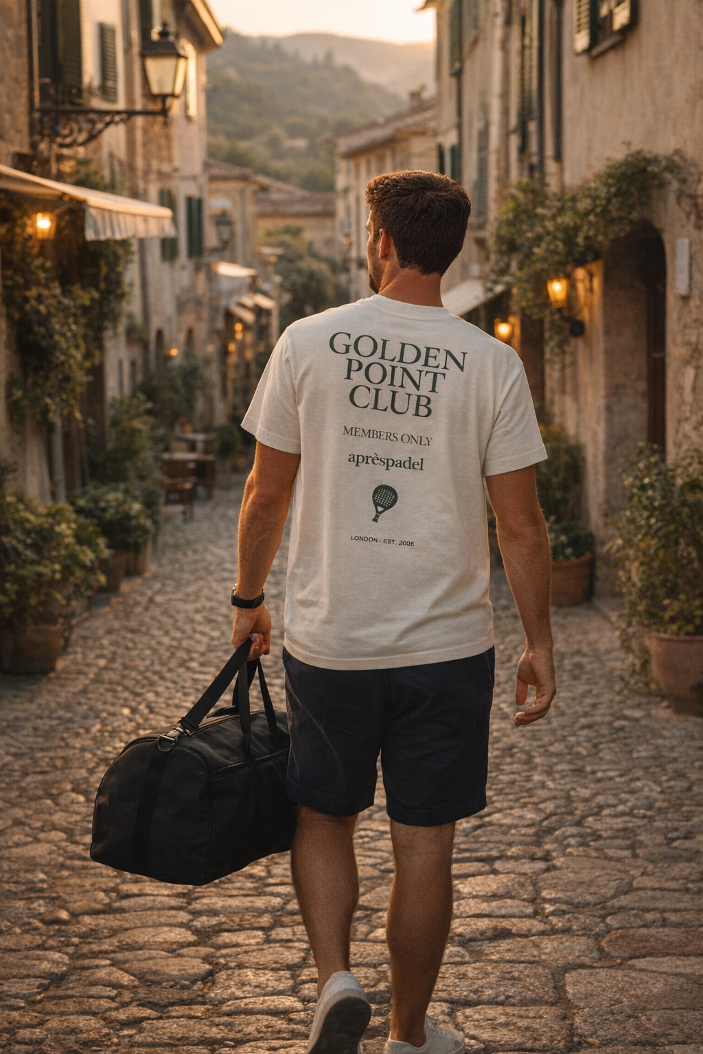 Man walking down a cobblestone street in an Italian town wearing a 'Golden Point Club' t-shirt wearing the AP Club Golden Point club tshirt