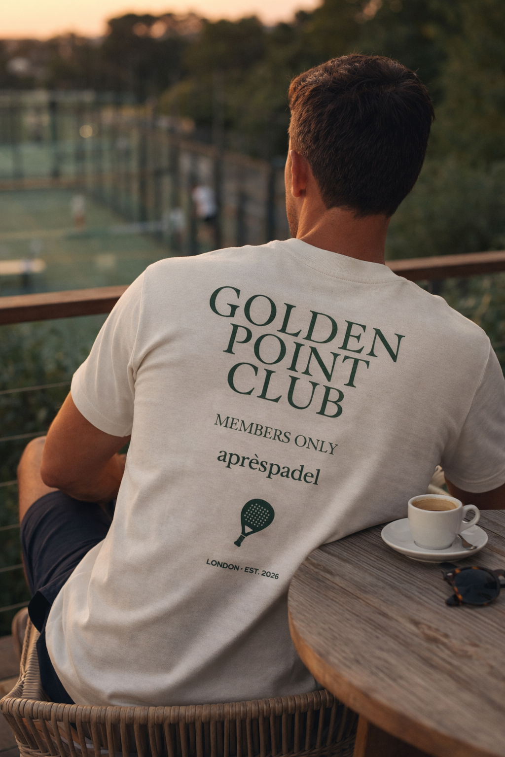 Man wearing a 'Golden Point Club' t-shirt sitting at a table with a cup of coffee, looking over a padel court.