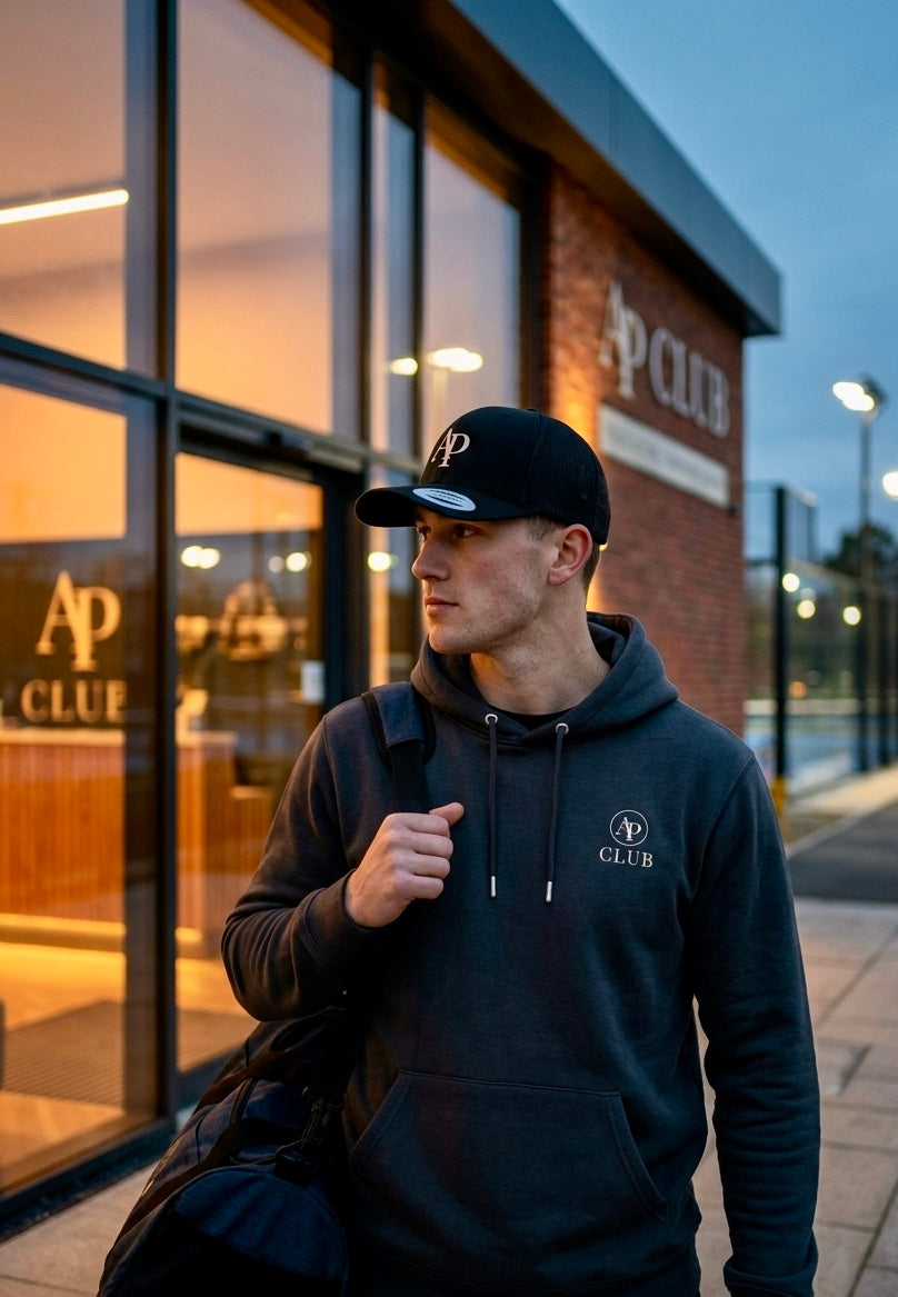 Man wearing a grey AP Club hoodie and AP Club cap with 'AP Club' branding in front of a padel building.