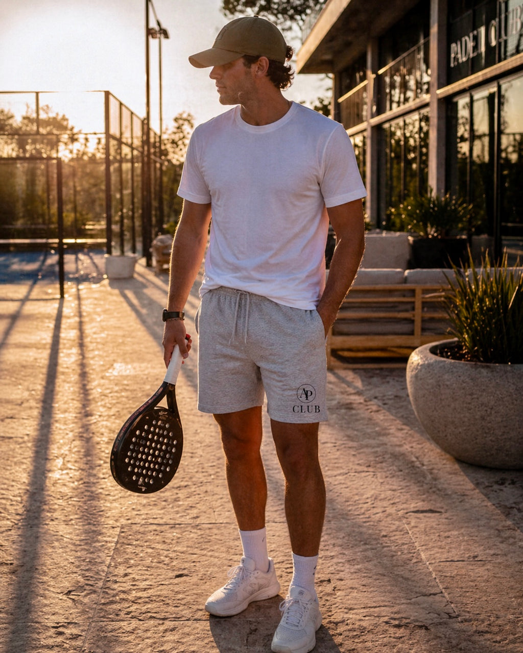 Man holding a paddle on a padel court with a building and plants in the background wearing AP Club shorts