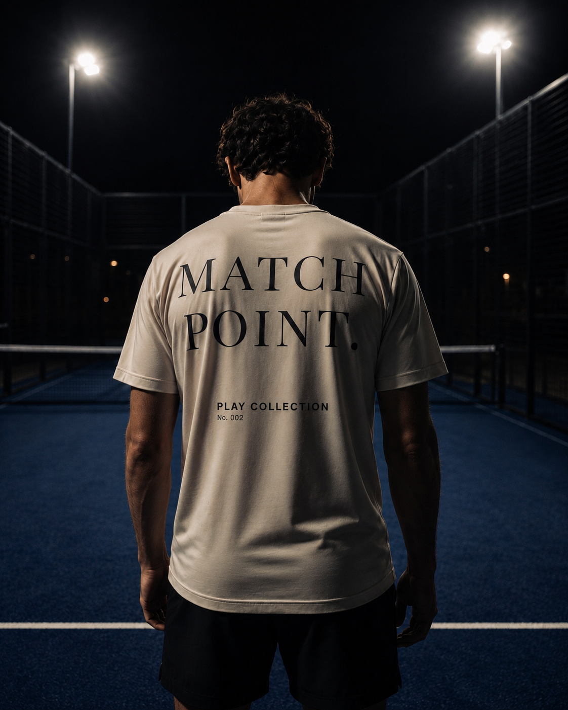 A man stands on a padel court at night, viewed from behind, wearing a light beige AP Club padel t-shirt with 'MATCH POINT PLAY COLLECTION' printed in black across the back.