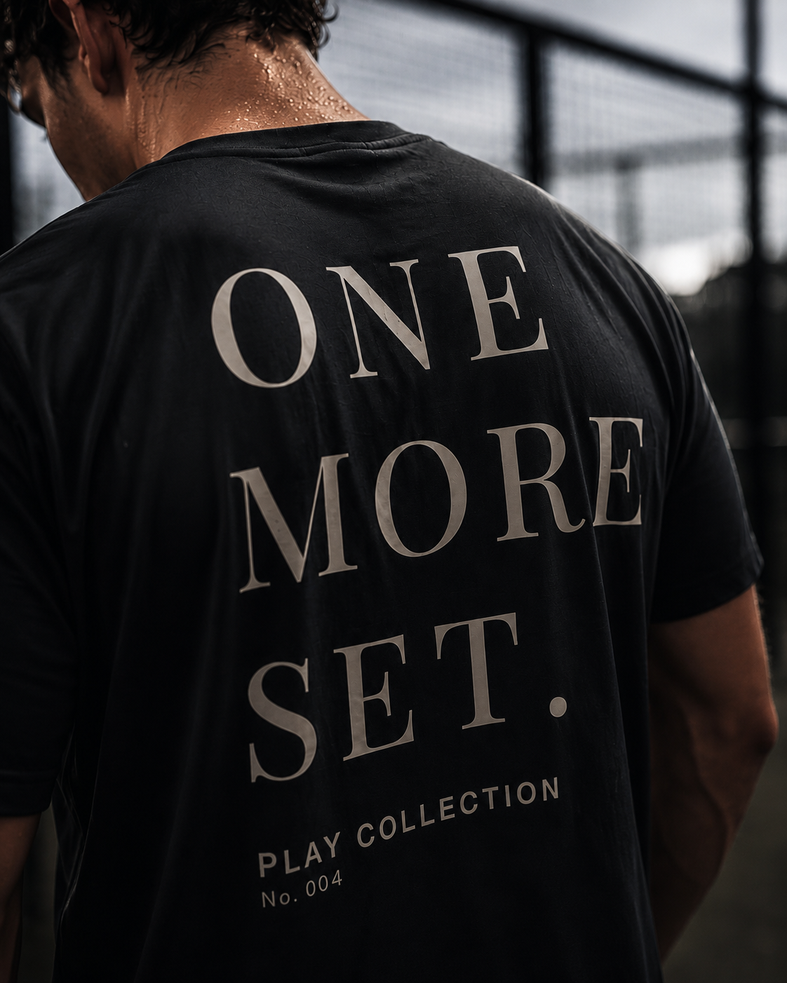 A padel player viewed from behind wearing a charcoal athletic AP Club padel t-shirt with large beige text reading 'ONE MORE SET.' and smaller text 'PLAY COLLECTION No.