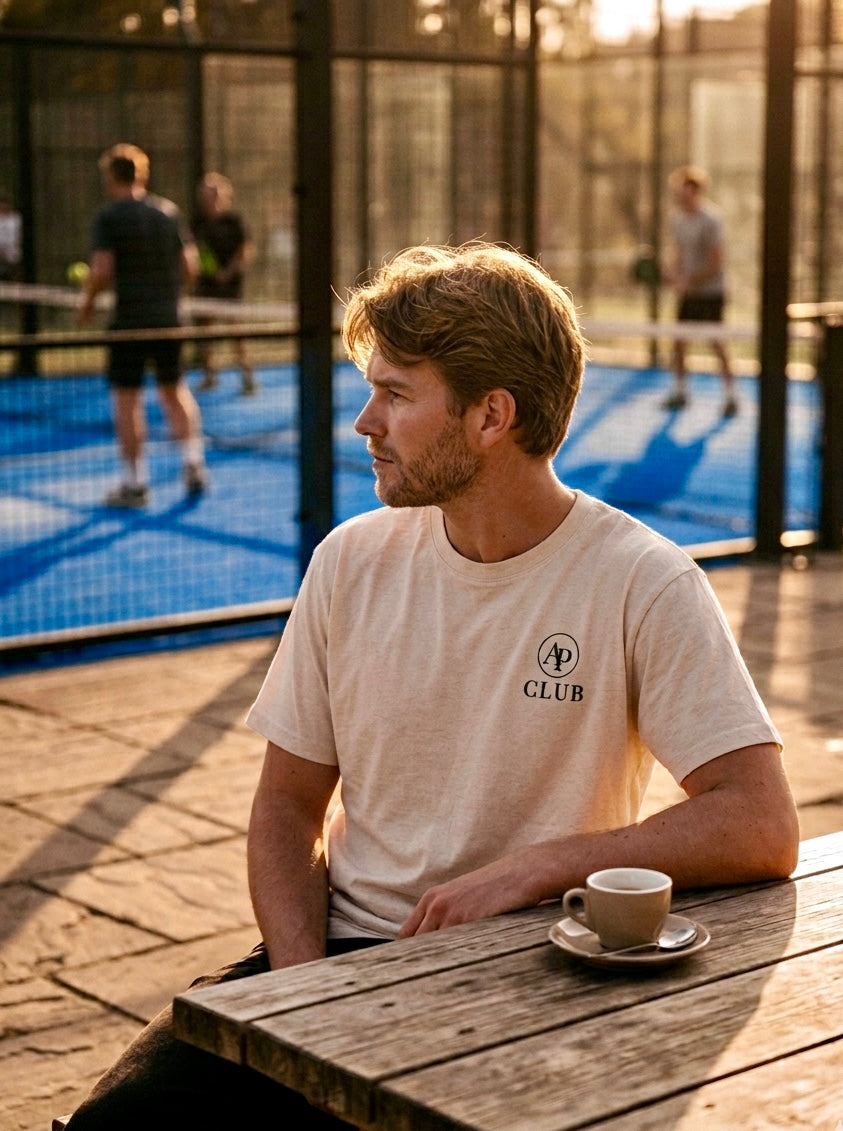Man sitting at a wooden table with a cup of coffee, looking towards a padel court wearing a cream AP Club padel tshirt