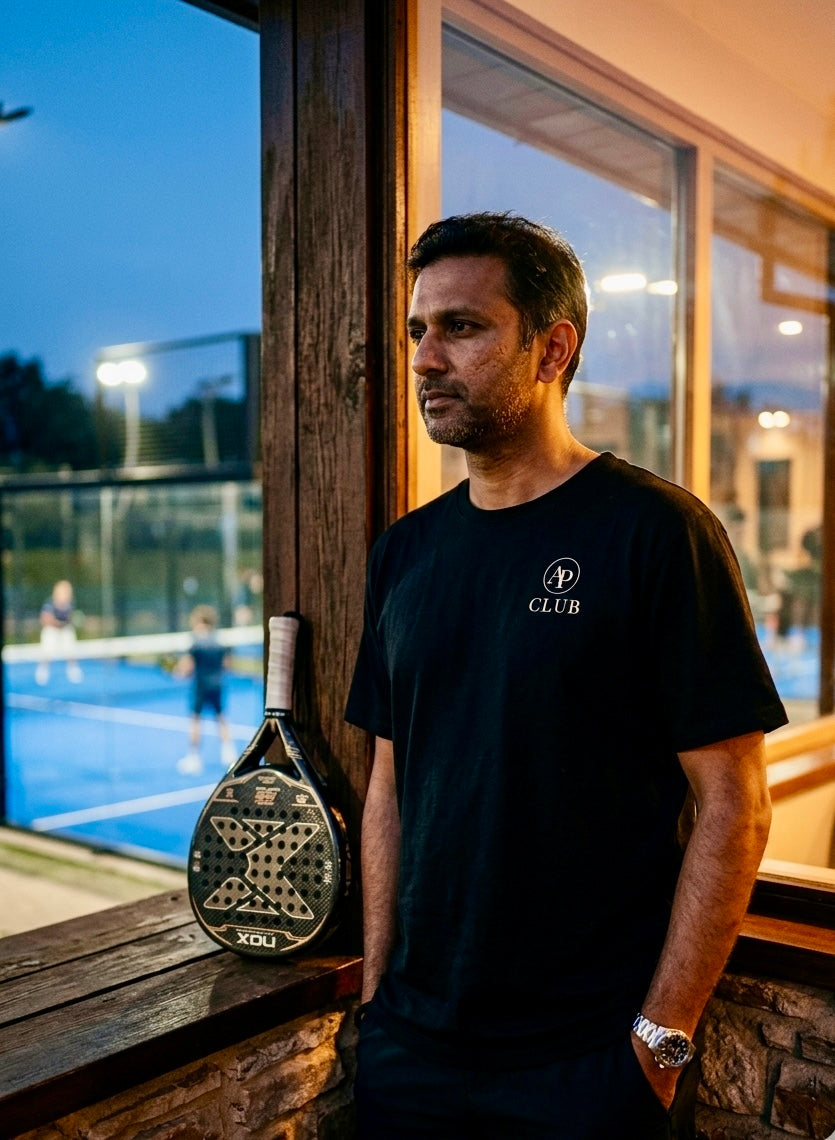 Man holding a paddle racket leaning against a window with an outdoor padel court visible, wearing a black casual AP Club padel tshirt