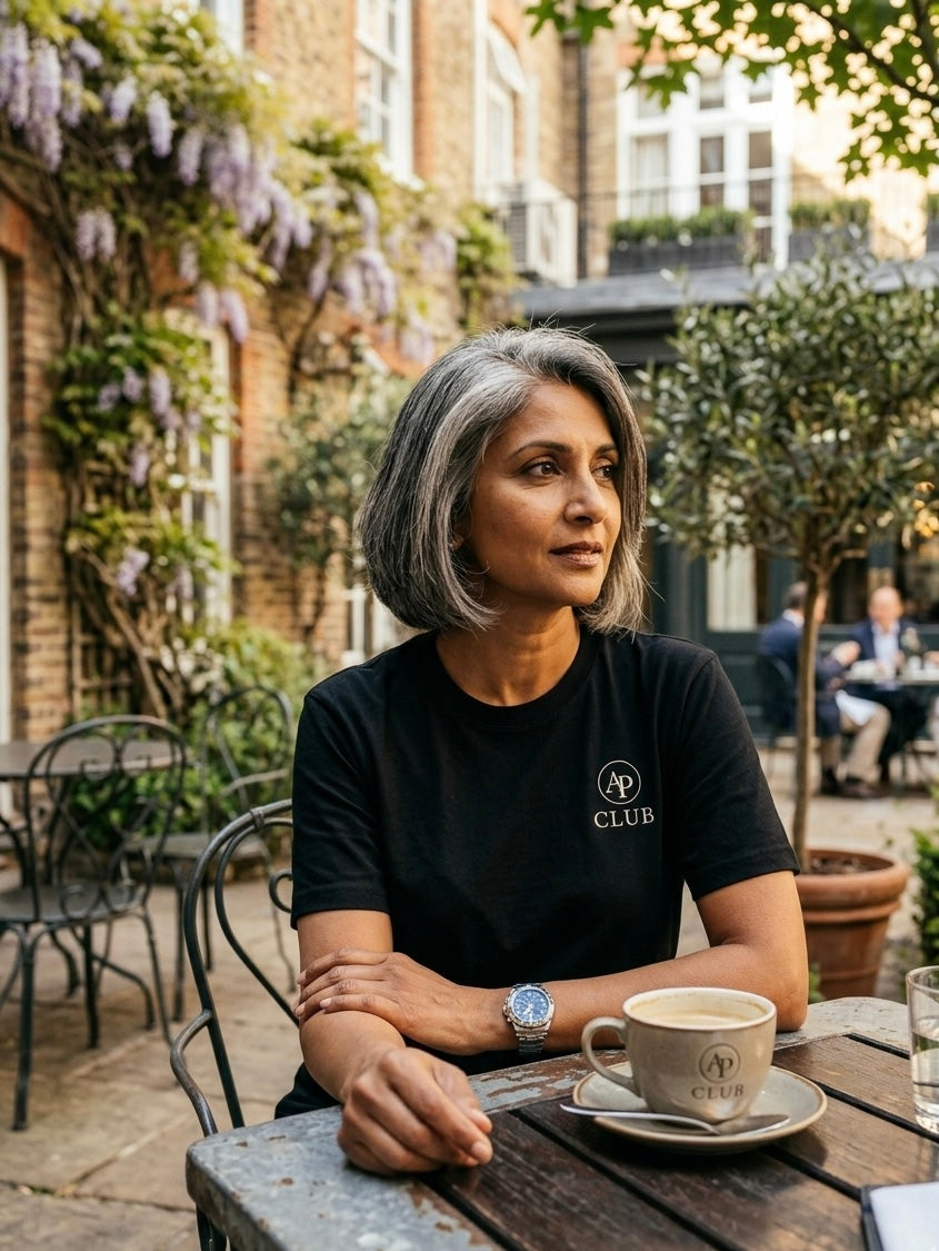 Woman sitting at an outdoor cafe table with a cup of coffee, surrounded by greenery and buildings wearing a black AP Club padel tshirt