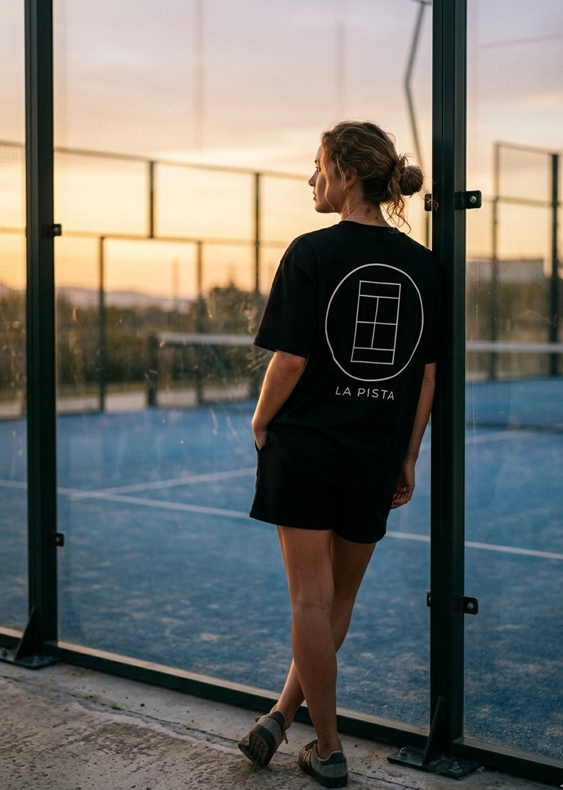 Person standing on a padel court with 'La Pista' logo on a black AP Club t-shirt, sunset in the background.