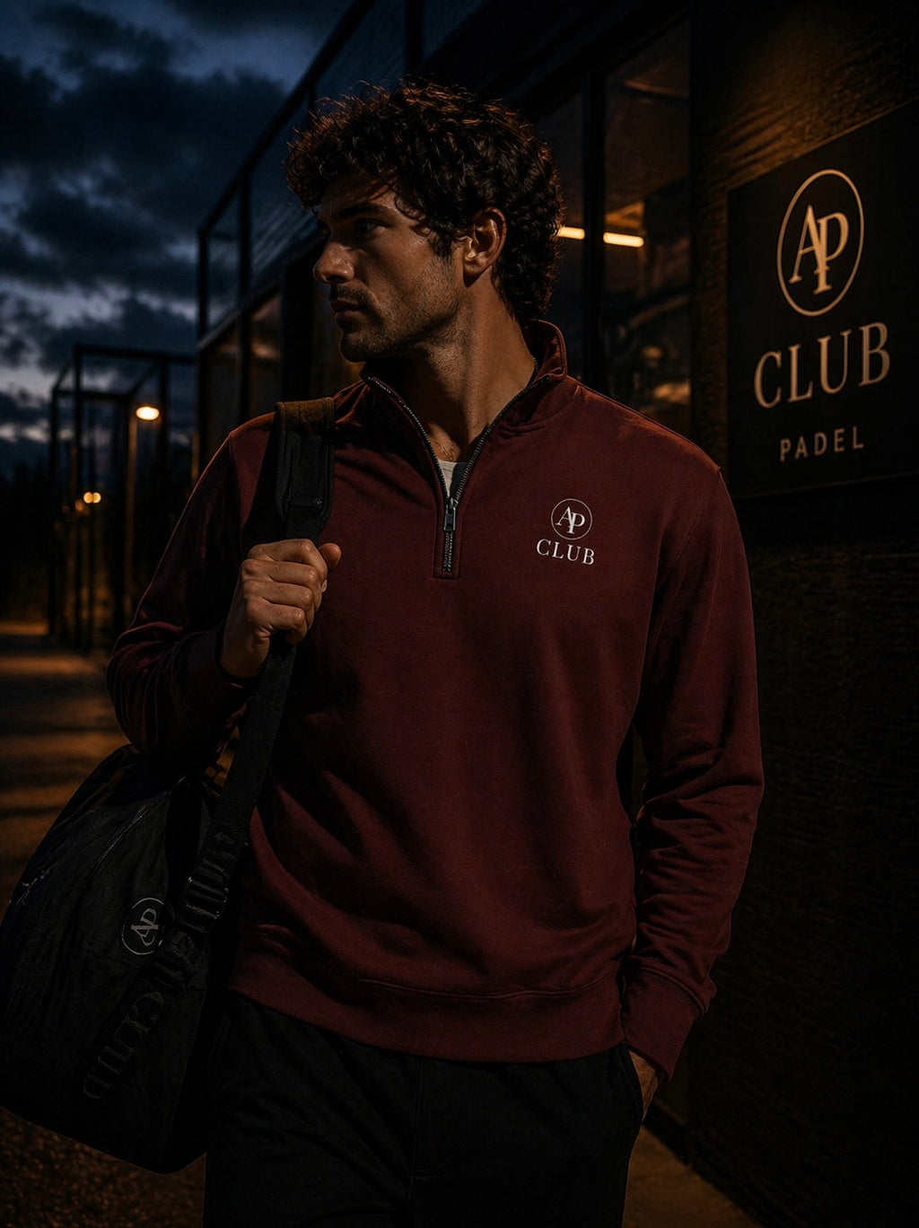 Man wearing a maroon AP Club Quarter Zip with a logo, holding a bag, standing in front of a building with 'AP Club' branding.