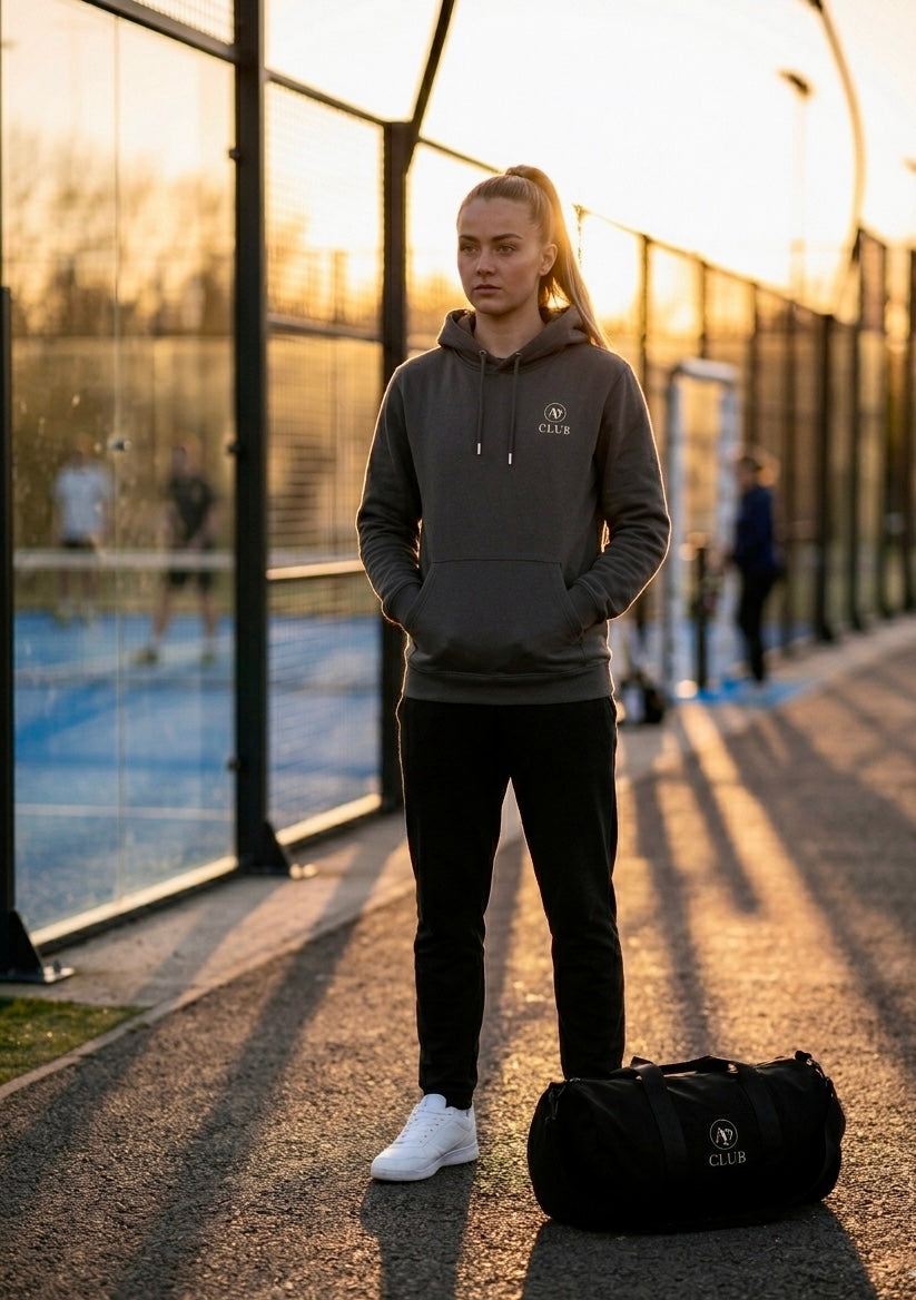 Person wearing an AP Club Vamos hoodie and black pants standing on a path with an AP Club duffel bag nearby.
