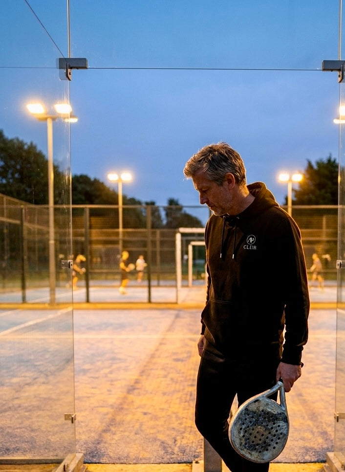 Man holding a padel on a padel court at dusk wearing a black AP Club Vamos hoodie
